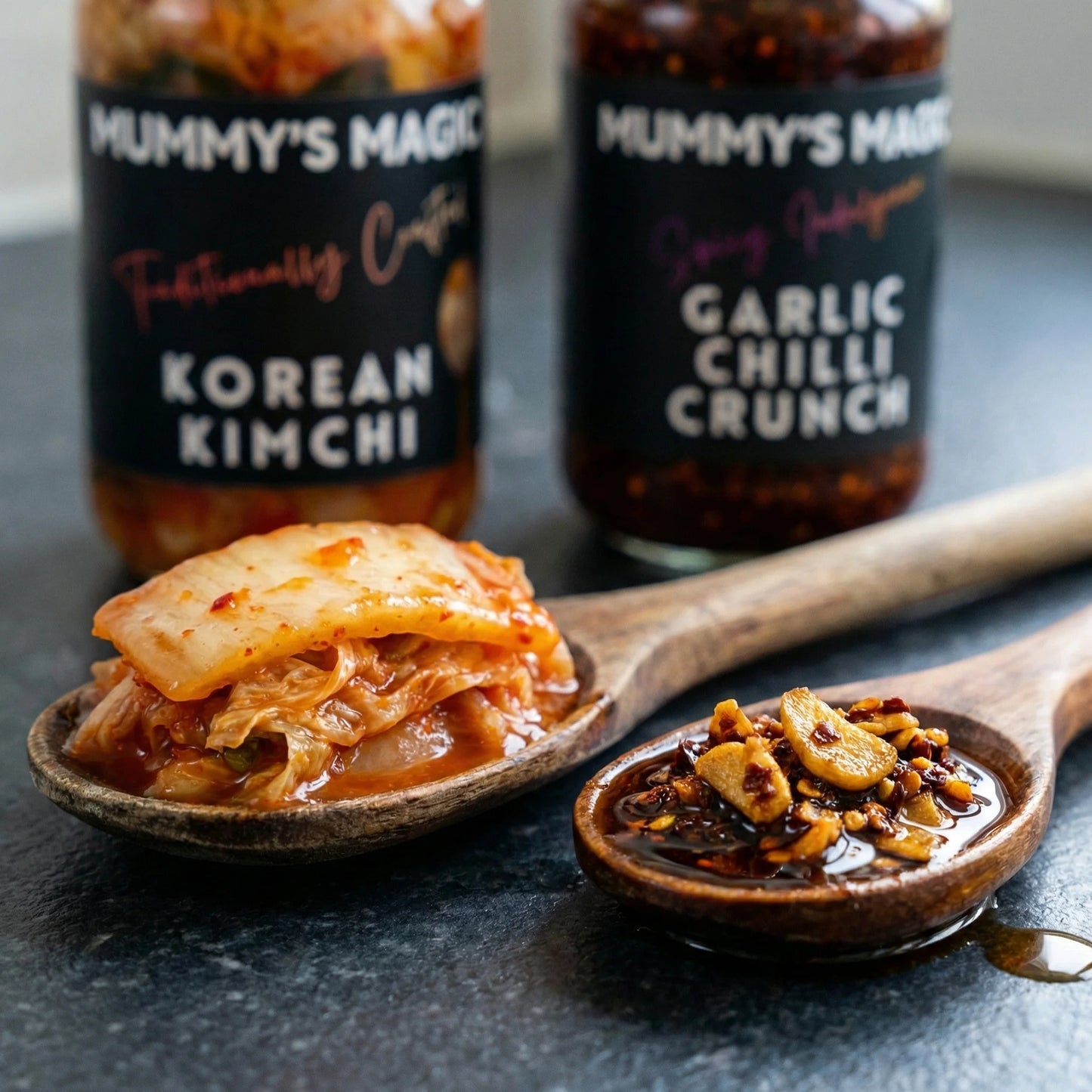 Two bottles of 'Mummy's Magic' food products with a wooden spoonful of Korean Kimchi and Garlic Chilli Crunch on a dark surface.